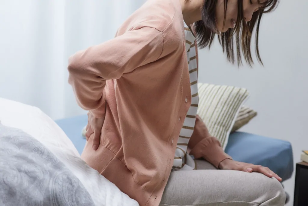Woman sitting on the edge of her bed holding her lower back in discomfort