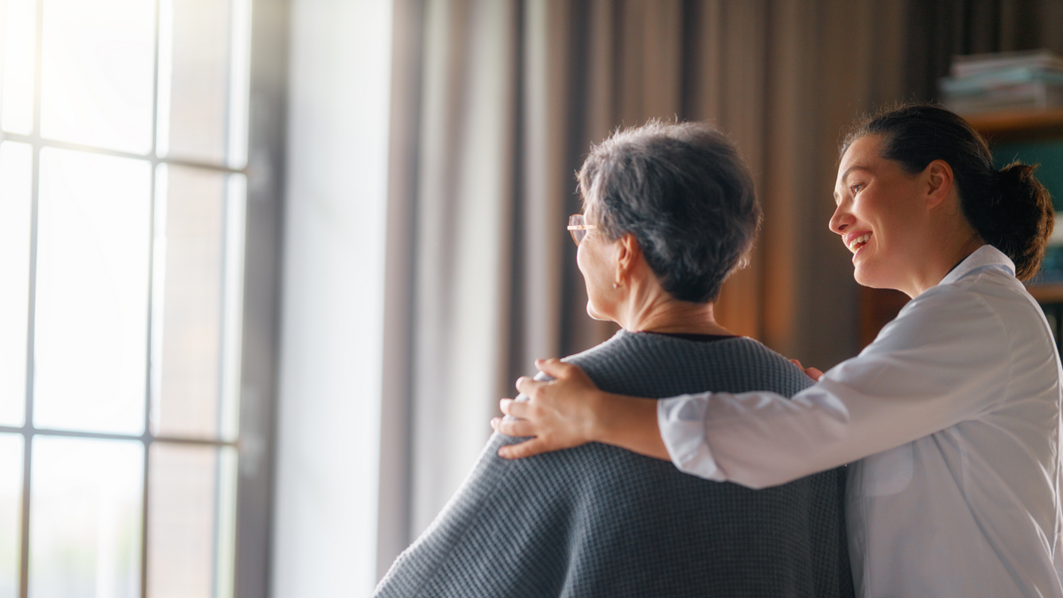 Caregiver with her arm around an older woman as they look out a window at home
